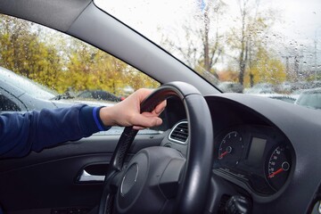 The driver's hand is on the steering wheel in the car, there are drops of autumn rain on the windows