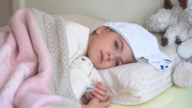 Sick fever cough child lying in bed with cold compress on his forehead, measure body temperature with electronic thermometer.Children cold and flu, coronavirus illness concept.