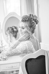 A young beautiful bride with a wedding hairstyle and makeup in the morning in a floor-length white lace tunic near the boudoir table. wedding morning