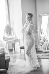 A young beautiful bride with a wedding hairstyle and makeup in the morning in a floor-length white lace tunic near the boudoir table. wedding morning