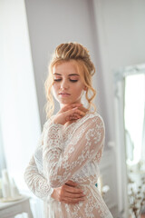 A young beautiful bride with a wedding hairstyle and makeup in the morning in a floor-length white lace tunic near the boudoir table. wedding morning