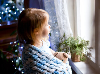 Cute little blonde girl wrapped in a plaid hugging a toy teddy bear looking out of window. Christmas lights sparkle in the background