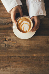 Female hands holding white cup with cappuccino on wooden background.