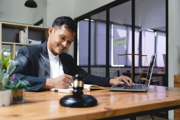 Professional Asian businessman, lawyer or financial consultant in formal suit working at his office desk, using laptop and taking notes on his book