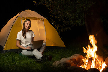Young freelance woman working on a laptop in the evening. A female tourist is sitting by a campfire and a tent