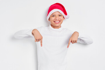 Portrait of smiling Asian man in christmas hat pointing fingers down, inviting people to follow...