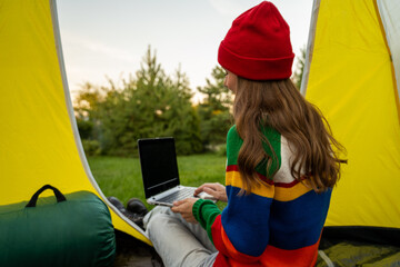 A cheerful woman in a red hat, relaxing in nature, is sitting in her tent with a laptop. 