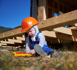 Boy toddler playing as builder on construction site. Child carpenter in orange helmet and blue overalls learning to build wooden frame house outdoor on sunny day. Carpentry and workshop concept.