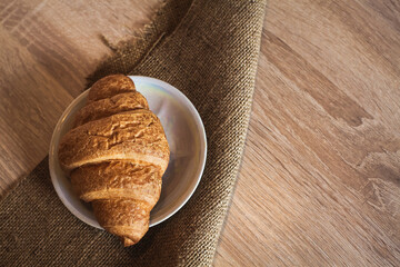An appetizing croissant lies in a saucer on a fabric, on a wooden light brown textured table.