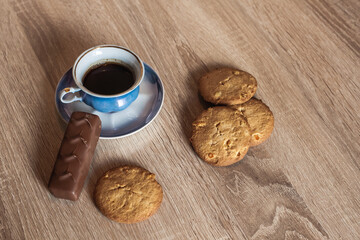 A cup of hot fragrant black coffee in a saucer with cookies and a chocolate bar, on a wooden light brown textured table.
