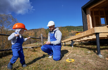 Father with toddler son building wooden frame house. Boy helping his daddy, playing with tape measure on construction site, wearing helmet and blue overalls on sunny day. Carpentry and family concept.