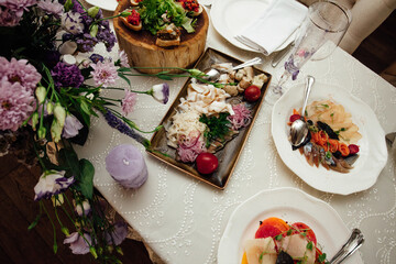Festive banquet served table. On it are candles, glasses, vegetables, meat, fish, herbs, sandwiches, tomatoes, onions in plates. Nearby is a vase with a bouquet of flowers.