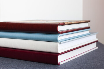 Four stylish photo books with a leather cover, white, burgundy, brown and blue, of different thicknesses, lie on a gray tabletop, against a light wall, indoors.