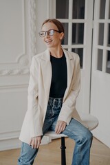 Happy pretty businesswoman in glasses sitting on little round table, posing for photo, looking aside