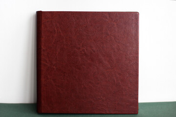 A large expensive premium photo book in a brown leather cover stands against a white wall on a green cloth indoors.