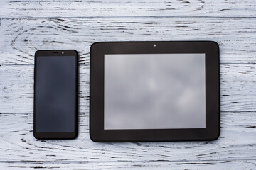Two black devices, a tablet and a mobile phone lie on a light wooden textured surface.