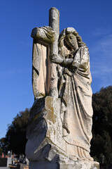 Worn and mossy stone sculpture of a woman wearing a robe while leaning on a crucifix in a cemetery, with bright blue sky in the background