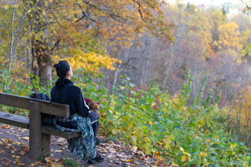 Fototapeta premium a young woman with a dog on her lap sits on a bench and observes the autumn landscape