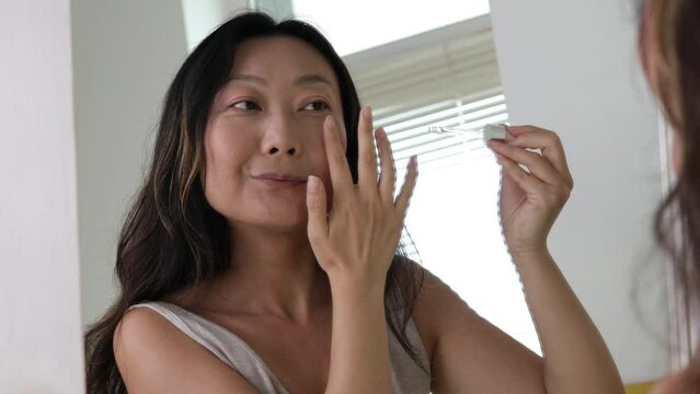 Skincare Concept. Beautiful Asian Lady Applying Face Serum With Dropper While Sitting In Front Of Mirror At Home, Smiling Woman Moisturizing Skin, Enjoying Self-Care Routine.