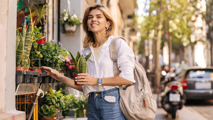 Pretty young caucasian girl holding pot with indoor flower standing outdoors in spring. Blonde wears casual clothes and backpack. Modern lifestyle concept