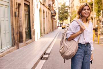 Pretty young caucasian woman goes to class with backpack along city street during day. Blonde wears shirt, jeans in warm weather. Urban life concept
