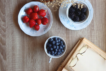 Vintage tea set filled with blueberries, blackberries and strawberries, open book, reading glasses and decorative flowers on the table. Flat lay.