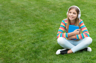 Happy teenage girl listening to audio training course sitting on grass after school, copy space