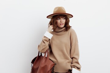 a delightful woman posing a white background in a sweater, shirt and hat gently touches her hair while holding a bag on one hand