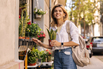 Nice caucasian young girl spends her time buying houseplant in open market. Blond woman wearing shirt with jeans looks towards European city. Lifestyle concept