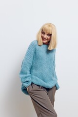  attractive woman in a beautiful light blue, long, knitted sweater stands on a light background in a relaxed pose and smiles pleasantly, turning her face to the side with her eyes closed