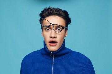 a handsome, attractive man stands on a light blue background in a blue zip-up sweater with black-rimmed glasses turned upside down on his face and looks in surprise at the camera with his mouth open
