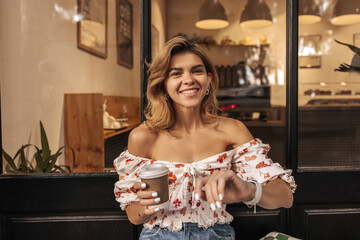 Cheerful european young lady smiling looking at camera enjoying thinning time drinking coffee outdoors. Blonde wears casual summer clothes. People emotions, lifestyle and fashion concept