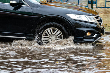 The car is driving through a puddle in heavy rain. Splashes of water from under the wheels of a car. Flooding and high water in the city.