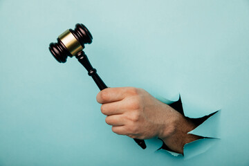 Hand holding a judge's gavel through torn blue paper wall. Law and courts, triumph of justice