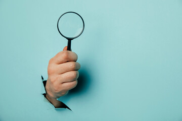 Male hand holds black magnifying glass in torn hole of blue background. Searching information concept