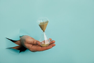 Male hand holds hpurglass in torn hole of blue background