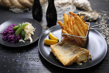 Fried fish fillets served with potato fries in a metal serving basket and salad mix, on black...