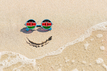 Fototapeta na wymiar A painted smile on the sand and sunglasses with the flag of the Kenya. The concept of a positive and successful holiday in the resort of the Kenya.