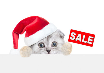 Scared tiny kitten wearing red christmas hat with sales symbol above empty white banner. isolated on white background