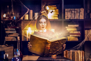 Surprised caucasian little girl in witch Cosplay reads magic book while holding magic wand,...