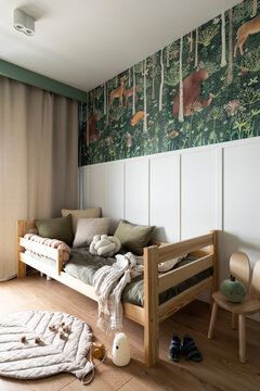 Creative composition of kids room interior with green forest wall paper, wooden bed, bottle green bedding, chair, simple gray curtain, rug and childrens accessories. Home decor. Template.