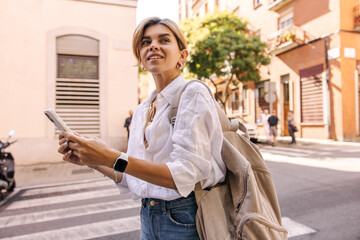 Attractive young caucasian woman is using internet outside standing on city street. Fair-haired lady wears casual clothes with backpack. Technology concept