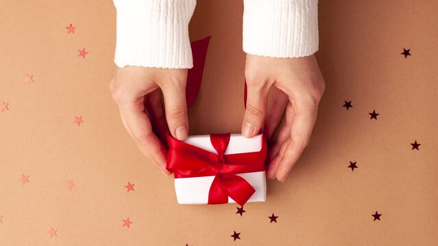 Human hands in white sweater hold a white paper gift with a red satin ribbon bow on brown background with red stars shapes. Stop motion animation Christmas Holidays concept flat lay.