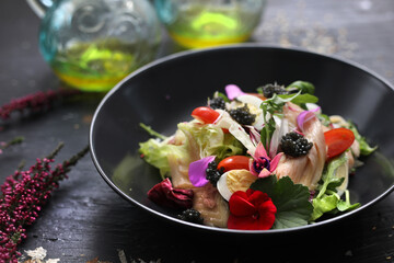 Salad with vegetables, fresh fish, caviar and quail egg, decorated with edible flowers. Salad in a black bowl, close up.