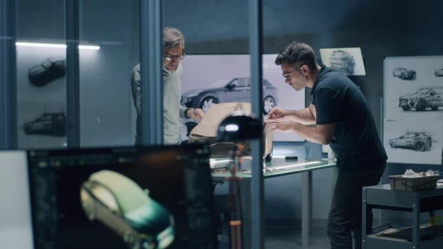 Two male automotive designers sculpting futuristic plasticine clay model of electric car with rake. Working in modern car design studio. Computer screen with software for 3D modeling projects.
