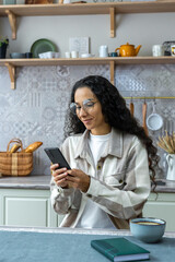 Vertical shot, beautiful hispanic woman using smartphone at home, woman with curly hair and glasses smiling, typing message and reading news online, student in kitchen.