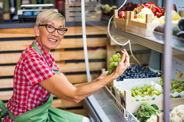 Woman works in fruits and vegetables shop. She is examining grapes.