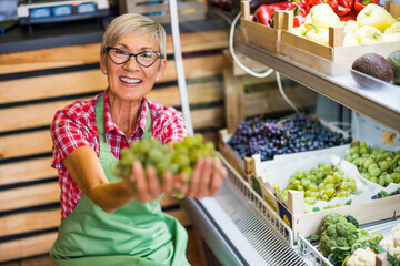 Woman works in fruits and vegetables shop. She is examining grapes.