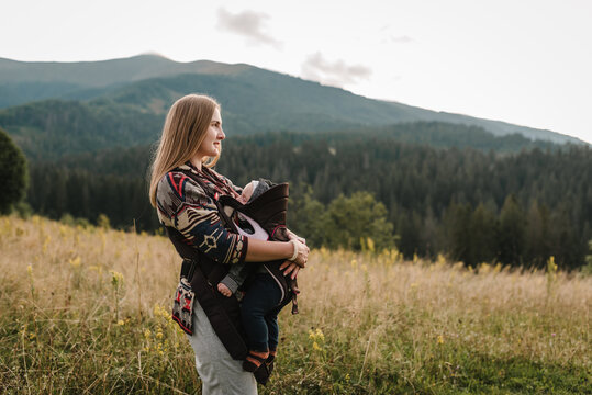 Carrying a baby. Mother with son in sling walk on orange grass in field. Mom, and child in autumn nature. Family with kid hiking. Young tourists on top of a mountain enjoying valley view sunset. - Powered by Adobe