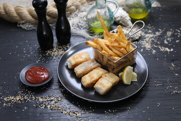Fried fish fillets served with potato fries in a metal serving basket, on a black plate, selective focus.